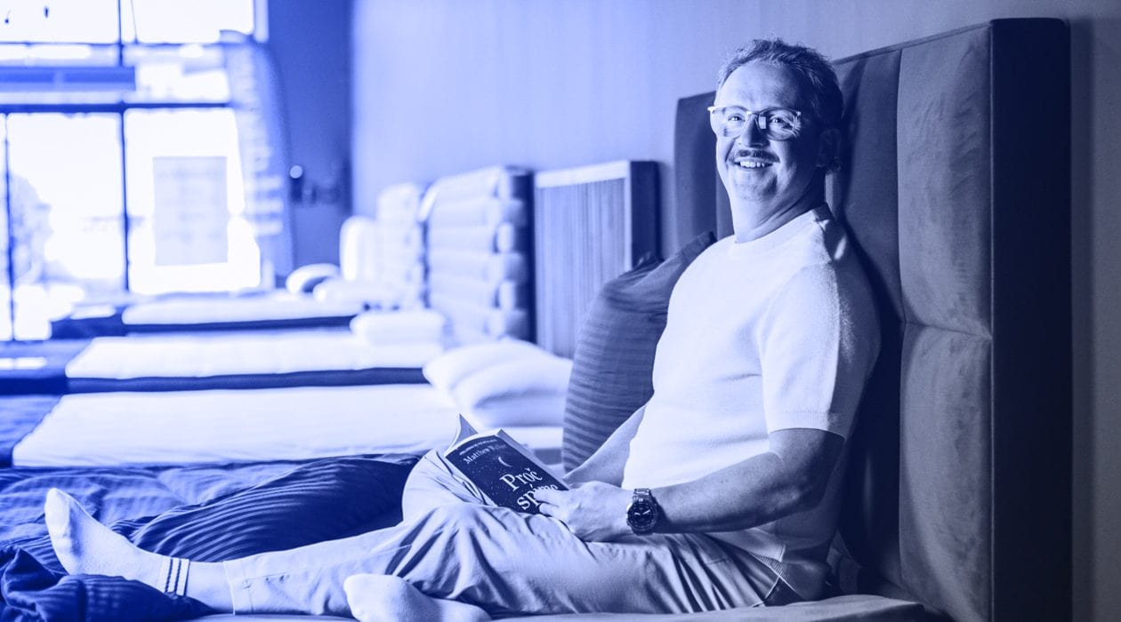 Vladan Hejnic sedí na posteli, čte knihu o lepším spánku, usmívá se, v moderním prostředí.