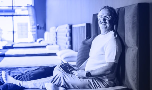 Vladan Hejnic sedí na posteli, čte knihu o lepším spánku, usmívá se, v moderním prostředí.